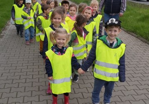 Dzieci ubrane w odblaskowe kamizelki idą parami chodnikiem.