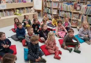 Dzieci siedzą na poduszkach na podłodze i słuchają opowieści na temat pracy bibliotekarza.