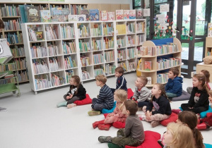 Dzieci siedzą na poduszkach na podłodze i słuchają czytanego przez Panią bibliotekarkę opowiadania.