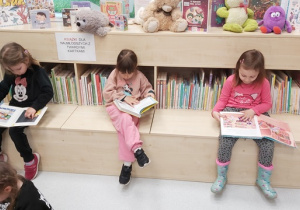 Trzy dziewczynki siedzą na siedzisku i oglądają książeczki dla dzieci.