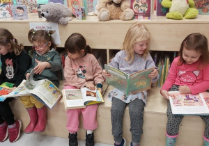 Dziewczynki siedzą na siedzisku, oglądają książeczki dla dzieci oraz komentują widziane ilustracje.