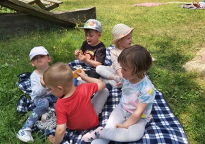 Dzieci z najmłodszej grupy siedząą na kocu i jedzą ciasteczka.