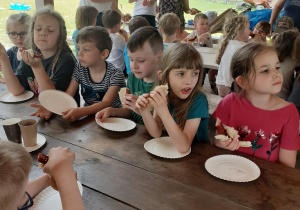 Dzieci siedzą przy stołach i jedzą pieczone kiełbaski.
