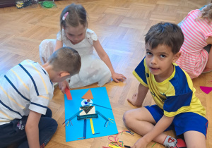 Troje dzieci siedzi na podłodze i wykonuje plakat do bajki Pinokio z przygotowanych materiałów plastycznych- kolorowy papie, patyczki,kolorowe sznurki