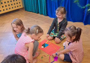 Cztery dziewczynki układają na dużym czerwonym kartonie elementy do wykonania plakatu.