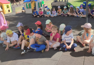 Dzieci z wsystkich grup siedzą na macie i słuchają poleceń prowadzącego zabawę.