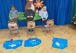 Troje dzieci stoi za krzesełkami z przyklejonymi sylwetami misia. W rękach trzymają sznurki nawinięte na rolkę jako wędki, którymi mają przyciągnąć złowione ryby.