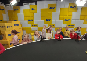Dzieci siedzą przy stole z mikrofonami na tle ścianki z napisami RMF FM.
