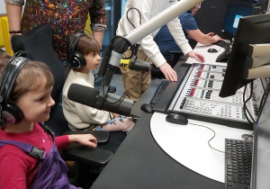 Dwie dziewczynki siedzą za pulpitem dźwiękowym z mikrofonami. Na uszach mają założone słuchawki.