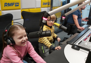 Dwoje dzieci siedzi za pulpitem dźwiękowym z mikrofonami. Na uszach mają założone słuchawki.