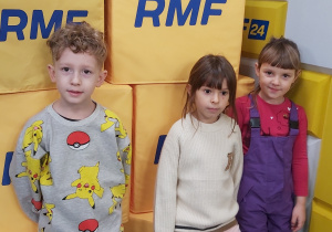 Troje dzieci stoi na tle pudeł z napisem RMF RM.