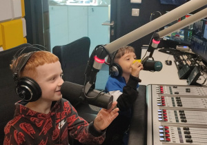 Dwóch chłopców siedzi za pulpitem dźwiękowym z mikrofonami. Na uszach mają założone słuchawki.