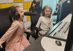 Dwie dziewczynki siedzą za pulpitem dźwiękowym z mikrofonami. Na uszach mają założone słuchawki. Jedna z nich mówi do mikrofonu.