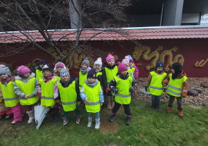 Grupa dzieci ubrana w odblaskowe kamizelki stoi przy murze ozdobionym smokami.