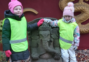 Dwie dziewczynki ubrane w odblaskowe kamizelki stoją przy rzeźbie przedstawiaącej słonia