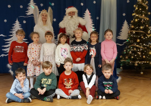 Dzieci z grupy 5 latków pozują do zdjęcia grupowego wraz ze Snieżynką i Mikołajem. Wszyscy stoją na tla zimowej dekoracji.