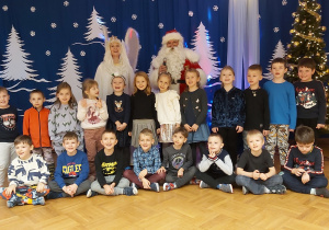 Dzieci z najstarszej grupy pozują do zdjęcia grupowego wraz ze Snieżynką i Mikołajem. Wszyscy stoją na tla zimowej dekoracji.