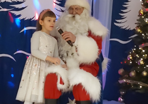 Dziewczynka stoi koło Mikołaja i śpiewa kolędę do mikrofonu.