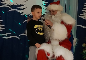 Chłopiec stoi koło Mikołaja i śpiewa kolędę do mikrofonu.