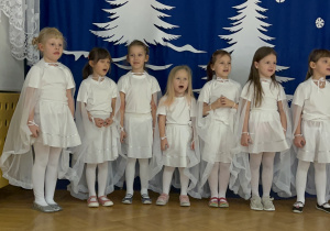 Dziewczynki stoją na tle zimowej dekoracji i śpiewają.