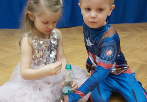 Dziewczynka przebrana za królewnę oraz chłopiec przebrany za policjanta biorą udział w konkursie- zbierają ziarenka fasoli do butelki.
