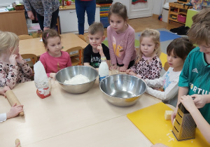 Dzieci siedzą przy stolikach, na których stoją dwie miski oraz mąka. Chłopiec