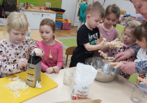 Dzieci przy stoliku przygotowują pizzę. Dwie dziewczynki ścierają na tarce ser żółty, natomiast kilkoro dzieci wyrabia rękami ciasto na pizzę.