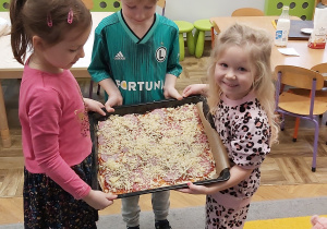 Dwie dziewczynki i chłopiec pokazują przygotowaną do pieczenia pizzę.