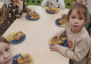 Dzieci siedzą przy stoliku, na którym znajdują się talerzyki z kawałkami upieczonej pizzy.