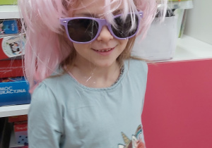 Dziewczynka w przebraniu. Na głowie ma różową perukę a na nosie okulary przeciwsłoneczne.