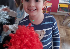 Uśmiechnięty chłopiec w pirackiej chustce trzyma w ręku biało-czerwoną perukę.