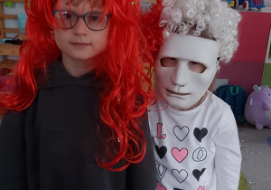 Dziewczynka z chłopcem pozują do zdęcia.Dziewczynka ubrana jest w biało-czerwonej perukę i maskę na twarzy. Chłopiec natomiast ma czerwoną perukę z długimi włosami.