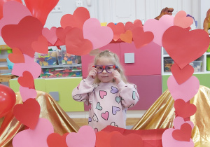 Dziewczynka pozuje do zdjęcia w fotoramce ozdobionej sercami i balonami w kształcie serca . Na nosie ma oprawki okularów ozdobione malutkimi serdszkami.