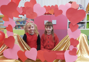 Dwie dziewczynki pozują do zdjęcia w fotoramce ozdobionej sercami i balonami w kształcie serca. Na nosie ma oprawki okularów ozdobione malutkimi serdszkami.