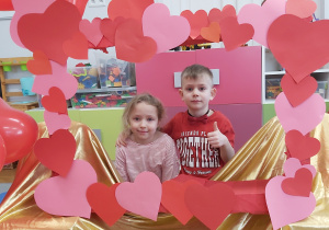 Dziewczynka i chłopiec pozują do zdjęcia w fotoramce ozdobionej sercamiz papieru i balonami w kształcie serca .