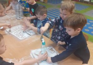 Dzieci za pomocą łyżki przesypują do butelki sól rozsypaną na stoliku.