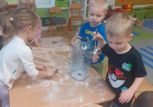 Dzieci za pomocą łyżki przesypują do dużej butelki sól rozsypaną na stoliku.