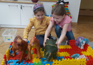 Dwie dziewczynki bawią się dinozaurami. Budują dla nich jaskinie z klocków.