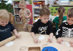 Dzieci przy stoliku wycinają z masy plastycznej dinozaury. Używają foremek w kształcie dinozaurów.