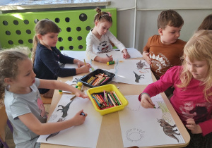 Dzieci wykonują pracę plastyczną przy stolikach. Nalepiają obrazki dinozaurów oraz uzupełniają w dowolny sposób obrazek.