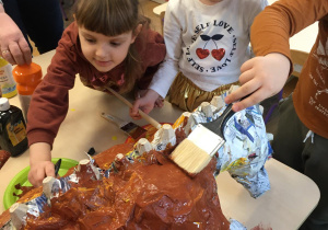 Dziewczynki malują zrobionego z gazet dinozaura brązową farbą.