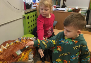 Dwoje dzieci maluje zrobionego z gazet dinozaura brązową i żółtą farbą.