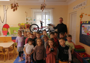 Dzieci stoją w grupie. Z tyłu stoi prowadzący zajęcia i trzyma rower.