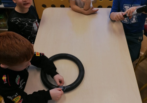 Dzieci siedzą przy stolikach. Jeden chłopiec trzyma dętkę. Dzieci uczą się jak załatać w niej dziurę .
