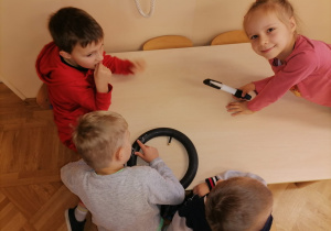 Dzieci siedzą przy stolikach. Jeden chłopiec trzyma dętkę. Dzieci uczą się jak załatać w niej dziurę .