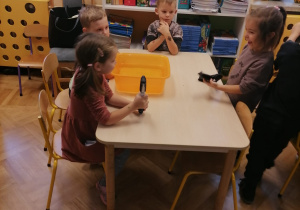 Dzieci siedzą przy stoliku, na którym stoi miska z wodą. Dzieci uczą się znajdować przy jej pomocy znajdować dziurę w dętce.
