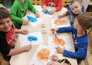 Dzieci siedzą przy stoliku. Rozmazują plastikowymi widelcami jednokolorowe plamy z farby.