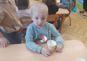 Chłopiec w szarej bluzce siedzi przy przy stoliku i trzyma w ręku kubek z wodą, w którym znajduje się słomka.