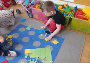 Chłopiec siedzi na podłodze i układa wymyślony przez siebie obrazek z wyciętych figur geometrycznych.