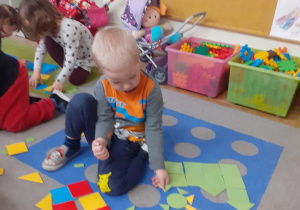 Chłopiec układa na dywanie obrazek z wyciętych figur geometrycznych.